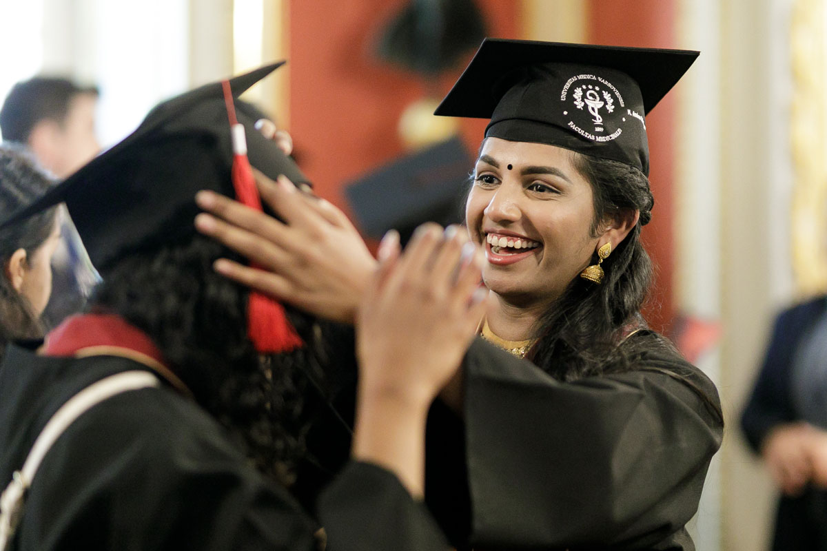 Two young women, one facing front, the other backwards. Dressed in black togas and black birettas. One of the girls has her hands next to the other girl's head, correcting her biretta.