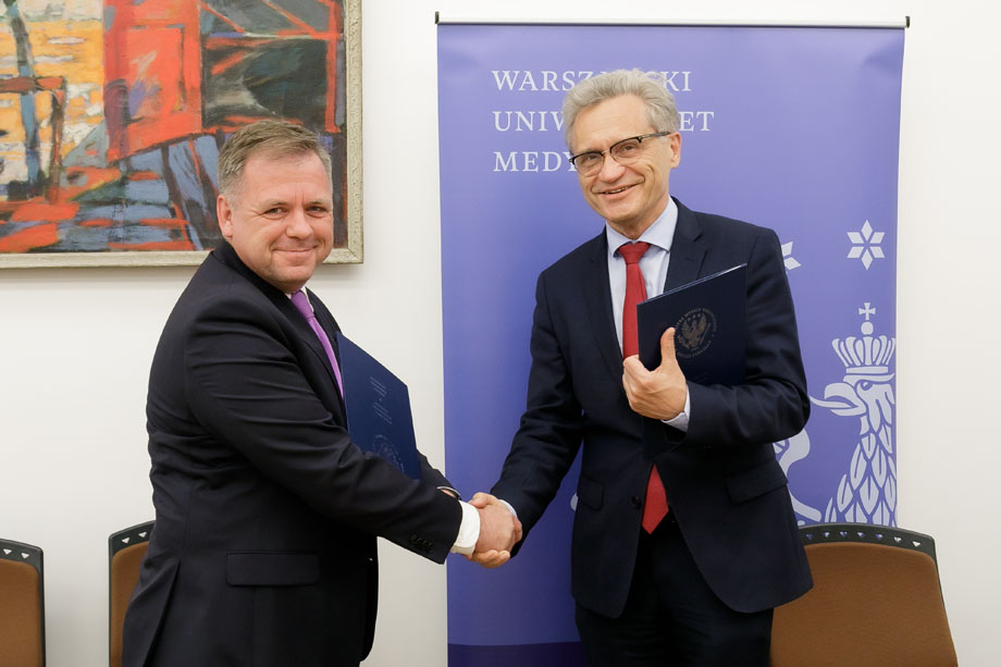 Two men in suits, shaking hands with their right hands, each holding a document folder in his left hand.