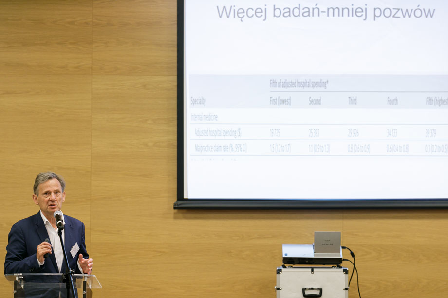 On the right, a man at the lectern. In the background, a screen showing the words "More research less lawsuits". 