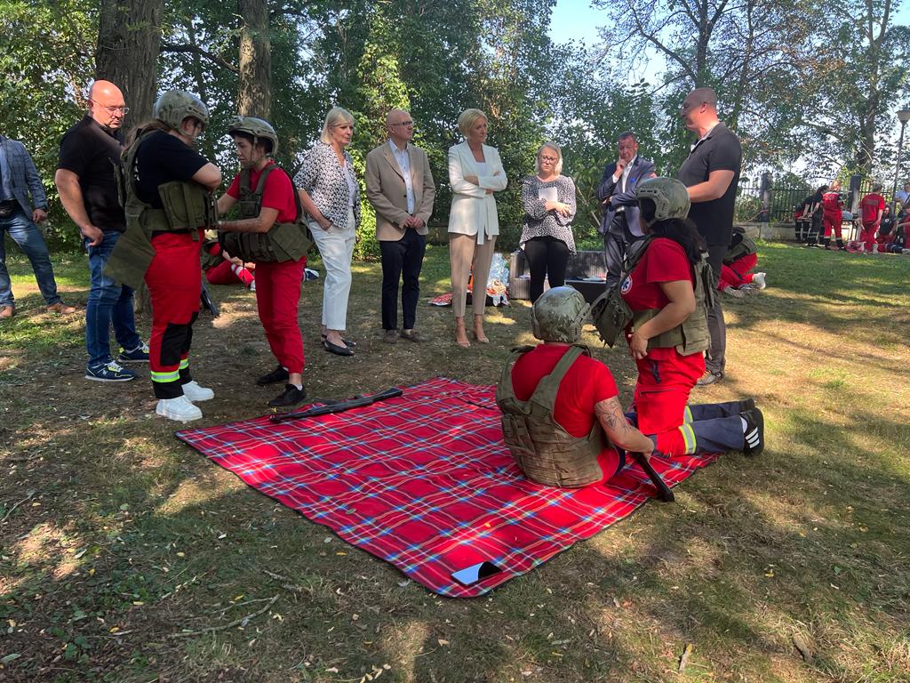 In the clearing a blanket is spread out. On it sits a woman wearing a helmet and a bulletproof vest. Next to her kneels another in the same outfit. There are several people standing around watching. Among them is a tall woman, blonde in a white jacket and beige pants.