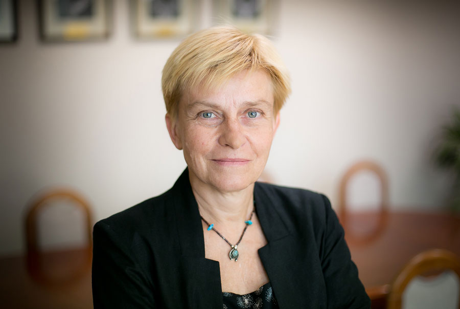 A woman with blonde hair, wearing a black jacket and a necklace smiles at the camera. In the background a brown table and chairs. 