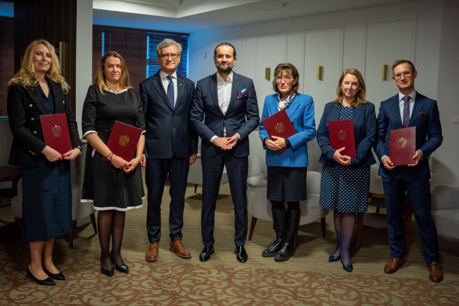 A group of seven people. Four women and three men. All are standing. Five with diplomas in hand. All elegantly dressed.