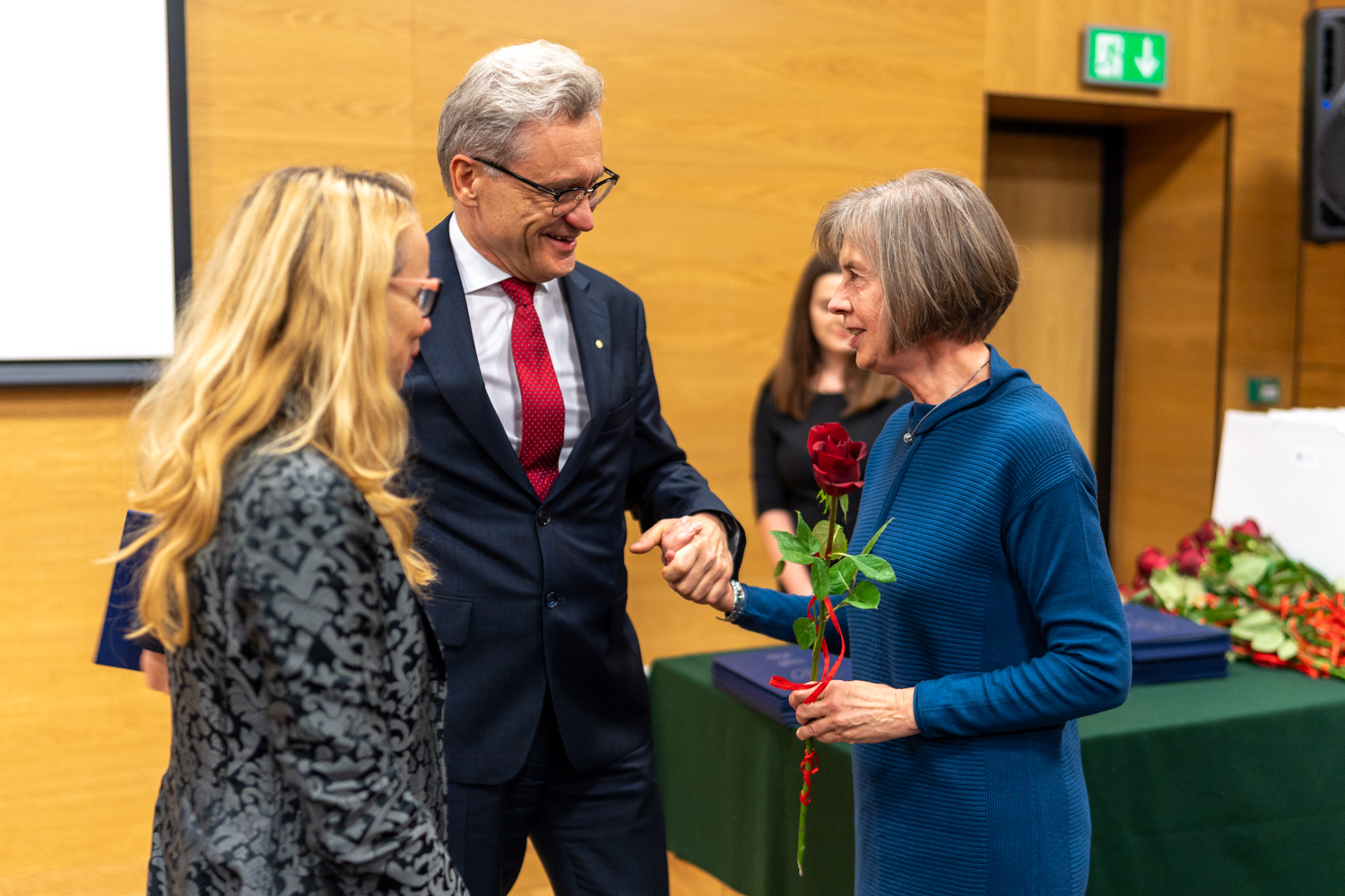 Starszy mężczyzna ściska rękę starszej kobiety i przekazuje jej dyplom. Bokiem stoi młodsza kobieta, blondynka.