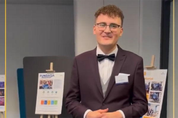 A young man in a suit and bow tie, wearing glasses, smiles. Behind his back you can see posters.