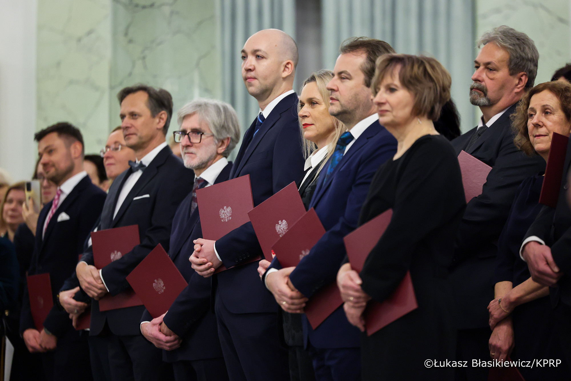 Prezydent wręczył nominacje profesorskie. Grono osób - nominowanych profesorów, stoją, w rękach trzymają teczki.