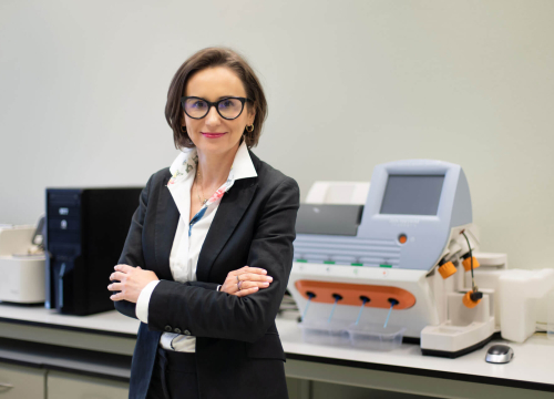 prof. Monika Czerwińska, uśmiechnięta kobieta, ubrana elegancko, stoi w pomieszczeniu laboratoryjnym