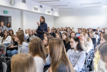 Mężczyzna - wiceminister stoi wśród grupy siedzących osób - studentów. Są w sali wykładowej.