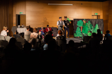 A group of people in a large hall. In the distance, actors on stage. At the back, a paper forest set. In the foreground, from the back, children and their parents