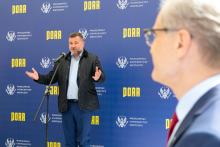 Middle-aged man with beard in suit, speaks into microphone, hands spread out