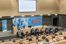 Sala konferencyjna. Widok z góry. Przy mównicy stoi mężczyzna w szarym garniturze. Za nim niebieski baner, powyżej ekran.