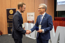 Rektor oraz student stoją na auli. Student odbiera nagrodę. Ściskają sobie dłonie. Stoją na auli konferencyjnej.