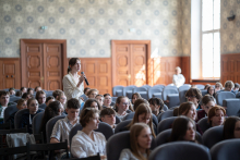 Aula. Młodzi ludzie siedzą w fotelach. Dziewczyna w białej bluzce stoi z mikrofonem i zadaje pytanie