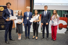 Grupa osób - studenci i rektor. Stoją w auli konferencyjnej. Studenci trzymają dyplomy. 