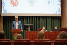 Mężczyzna w garniturze - rektor - stoi za mównicą i przemawia. Przed nim uczestnicy konferencji, za nim ekran multimedialny ze slajdem z konferencji.