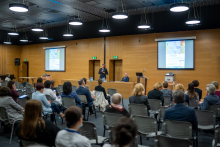 Sala konferencyjna, na krzesłach siedzą ludzie, widać ich od tyłu. W oddali stół, przy którym znajduje się dwóch mężczyzn, jeden stoi i mówi do mikrofonu. Drugi - siedzi.