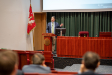 Sala konferencyjna, w oddali przy mównicy wykładowca gestykuluje.