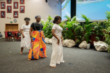 Three dark-skinned women dance lined up one behind the other. The first and last dressed in white. The middle one in a turban, black blouse and patterned skirt.