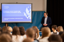 Lecture hall. In the foreground, heads of the audience visible from behind. At the end of the room a man in a suit and a headset microphone. In his hand he holds a remote control. Behind him on the screen you can see the title of the lecture and the logo of the Medical University of Warsaw.