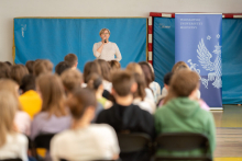 A gymnasium turned into a lecture hall. Audience visible from the back. At the end of the hall, a woman speaks into a microphone. 