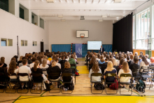 A gymnasium turned into a lecture hall. Audience visible from the back. Chairs occupied to the last seat. At the end of the room you can see the lecturer and the screen.