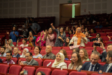 Audience. Many people sit in red seats. A few stand and ovate the performances.