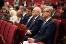 Audience. Many people are seated in red chairs. In the foreground are two older men in suits.