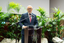 An elderly man at the lectern. Green palm trees in pots in the background.