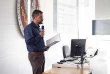 A man with a beard speaks into a microphone. In his hand he holds a briefcase. Next to him is a desk with a computer, behind him a white wall on which hangs the WUM emblem.