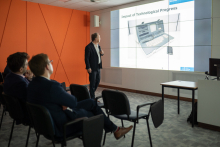 A standing man dressed in a suit. He looks at a multimedia screen with a presentation and speaks into a microphone. Behind him sit male lecture participants.