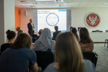 A standing man dressed in a suit. He looks at a multimedia screen with a presentation and speaks into a microphone. Behind him sits a group of various people, participants in the lecture. In the background is a white wall with the WUM emblem.