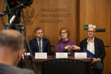 Stół konferencyjny. Siedzi dwóch mężczyzn oraz kobieta, która gestykulując mówi do mikrofonu.