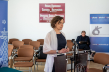 A woman in a dress and jumper, speaks behind the lectern. 