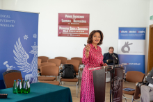 A woman in a pink dress, speaks behind the lectern, pointing to something with her hand. Behind her, a table and a rollup.