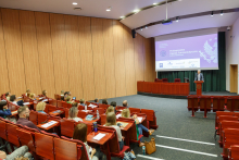 Aula wypełniona słuchaczami. W oddali na mównicy starszy mężczyzna w graniturze.