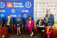 Po lewej starszy mężczyzna w szarym garniturze mówi do mikrofonu. Za nim siedzą prelegenci: dwóch mężczyzn i dwie kobiety. Za nimi na ścianie plakat z napisami Szczyt Zdrowie.