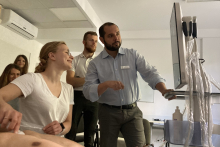 The lecturer - a middle-aged man with a beard - stands at the ultrasound machine. Next to him sits a young girl - a student is performing an ultrasound. Behind them other students.