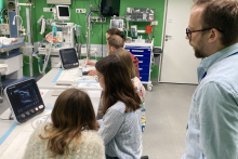 A room like a hospital room with specialized equipment. A long table at which five students sit . On the table two ultrasound machines. 