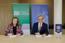 A woman in glasses and a checked jacket and a man in glasses and a suit sit behind a table. Documents lie in front of them. Both are holding pens in their hands.