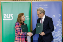 On the left, a woman wearing glasses and a checked jacket. On the right, a man in glasses and a suit. They stand and shake hands, looking at each other. In their hands they hold green briefcases.