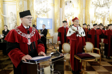 Grupa osób w bogato zdobionej sali zamkowej. Na pierwszym planie mężczyzna w czerwono-czarnej todze, stoi za mównicą i przemawia. Ma czarny biret i okulary. W oddali grupa osób ubranych w togi.