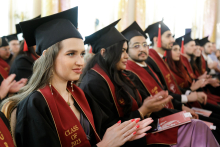 A group of seated young people dressed in black togas. They all have black birettas on their heads and maroon sashes slung around their necks. They are applauding.