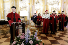 A group of people in the castle hall. On the left, a man in a red and black toga, stands behind the lectern and speaks. In front of the rostrum a bouquet of colourful flowers. On the right, a group of people in togas.