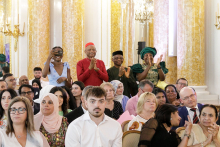 Group of people. Some are sitting. Four people stand and applaud. Dressed in different coloured outfits.