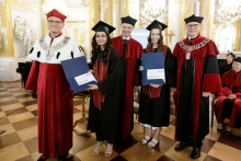 A group of five people standing. Three men and two young women. The women are standing in the middle. They hold gift bags and briefcases in their hands. 