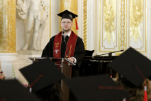 A young man wearing glasses, dressed in a black toga and biretta speaks behind the rostrum. 