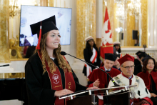 A young woman, dressed in a black toga and biretta, speaks behind the rostrum. In the background, several people dressed in red and black togas. The WUM banner can also be seen.