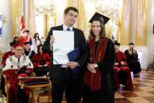 In the foreground, two standing people. On the left, a man in a suit and tie holding a diploma. On the right, a young woman wearing a biretta and a black toga. In the background sit people dressed in togas.