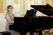 A woman in a beige dress sits behind a piano.