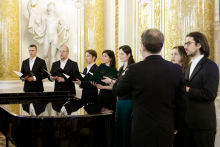 A group of people dressed in black, choristers. They are standing and singing. In their hands they hold briefcases.