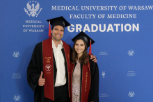 Two people, a young man and a young woman dressed in togas and birettas pose for a photo against a blue background with the WUM logo and information about the Diplomatatorium.